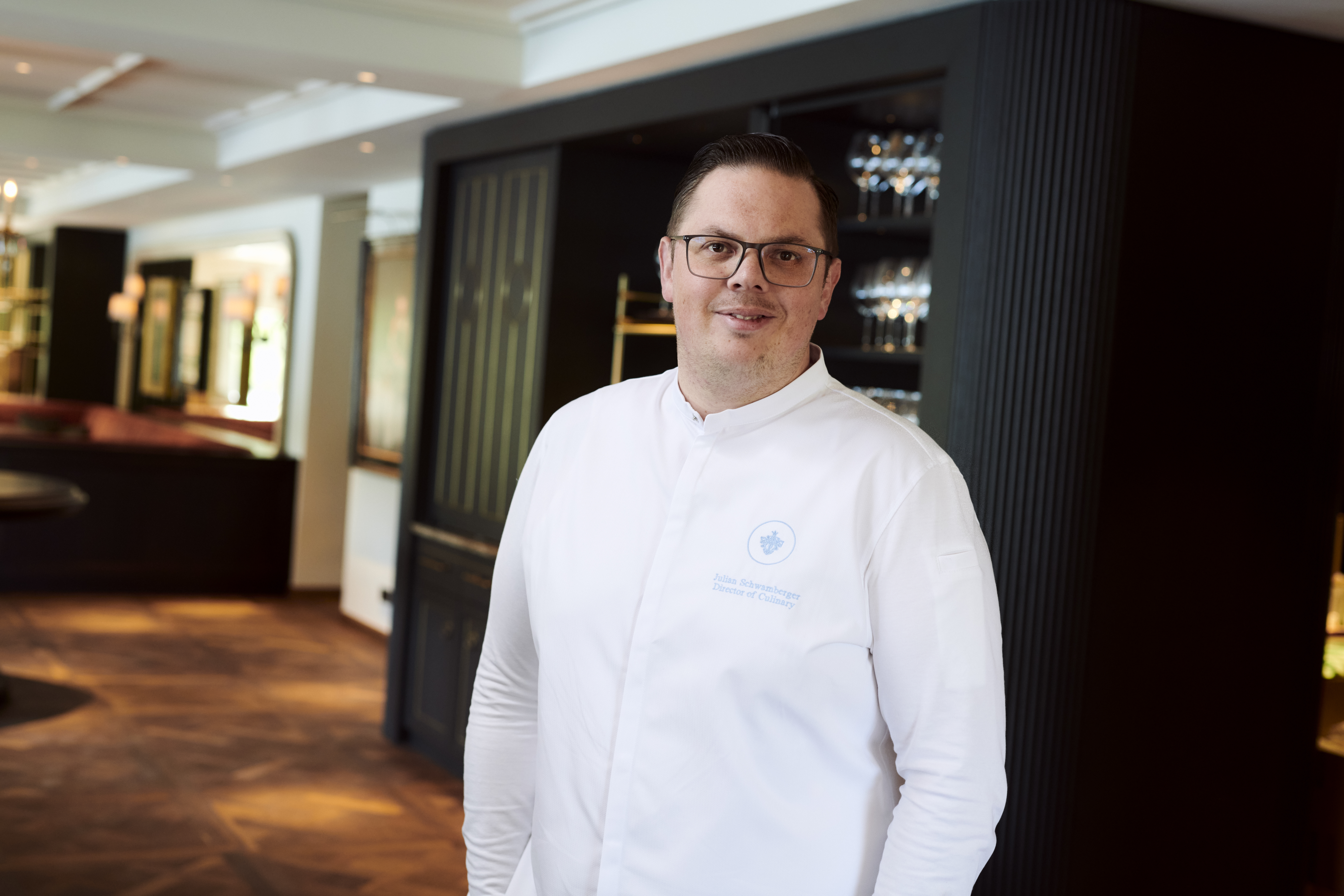 Smiling chef in white uniform standing inside an elegant restaurant interior.