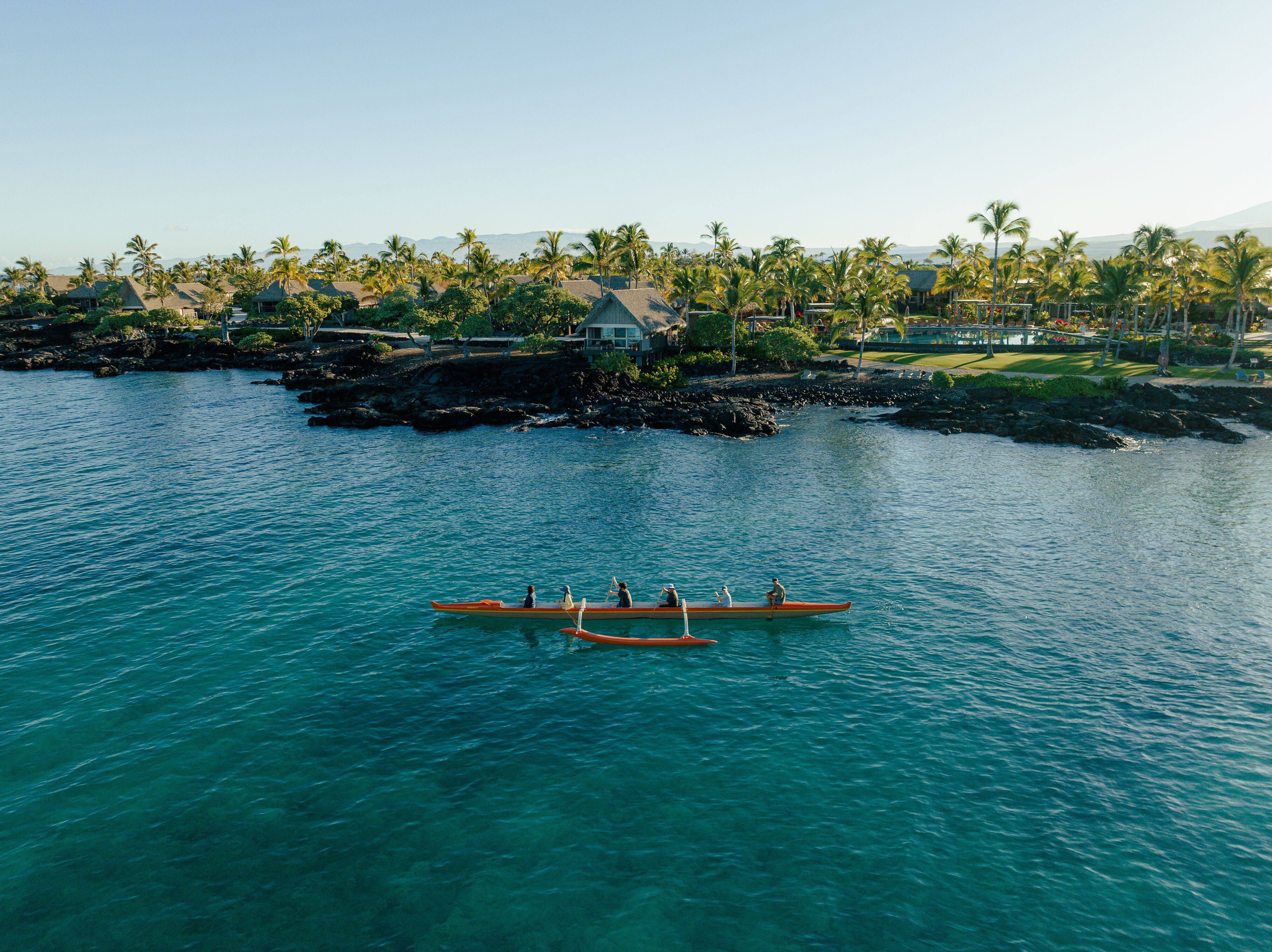 Six people paddling a canoe along the ocean coast with thatched roof bungalows in the background