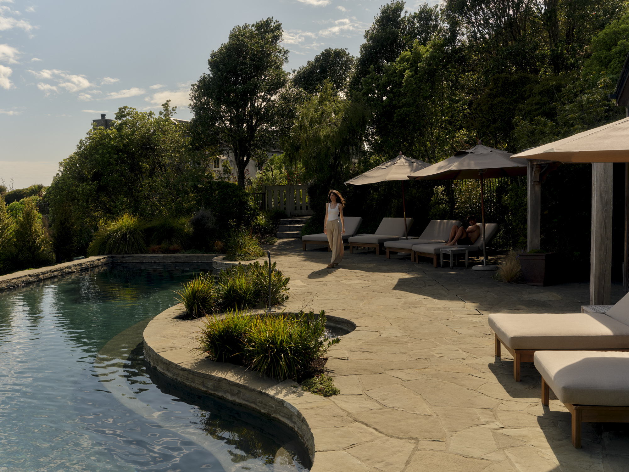 An outdoor pool surrounded by greenery with a wooden lodge, shaded lounge chairs, and a stone patio with lush plants.